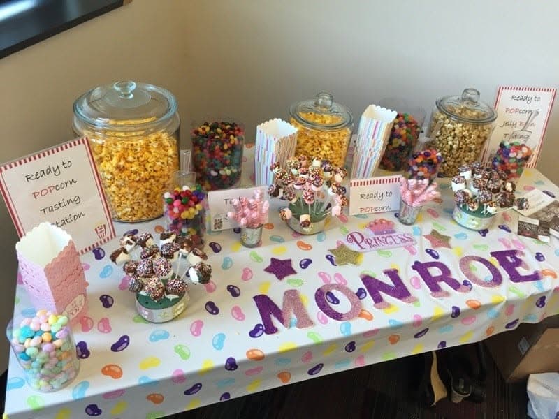 Colorful dessert table with popcorn jars, jelly beans, marshmallow pops, and glittery letters spelling Monroe.