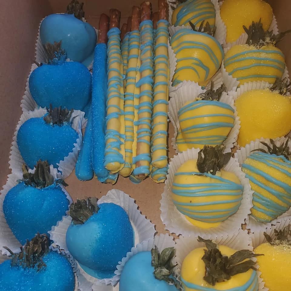 Assorted blue and yellow chocolate-covered strawberries and pretzel rods with decorative drizzles in a box.