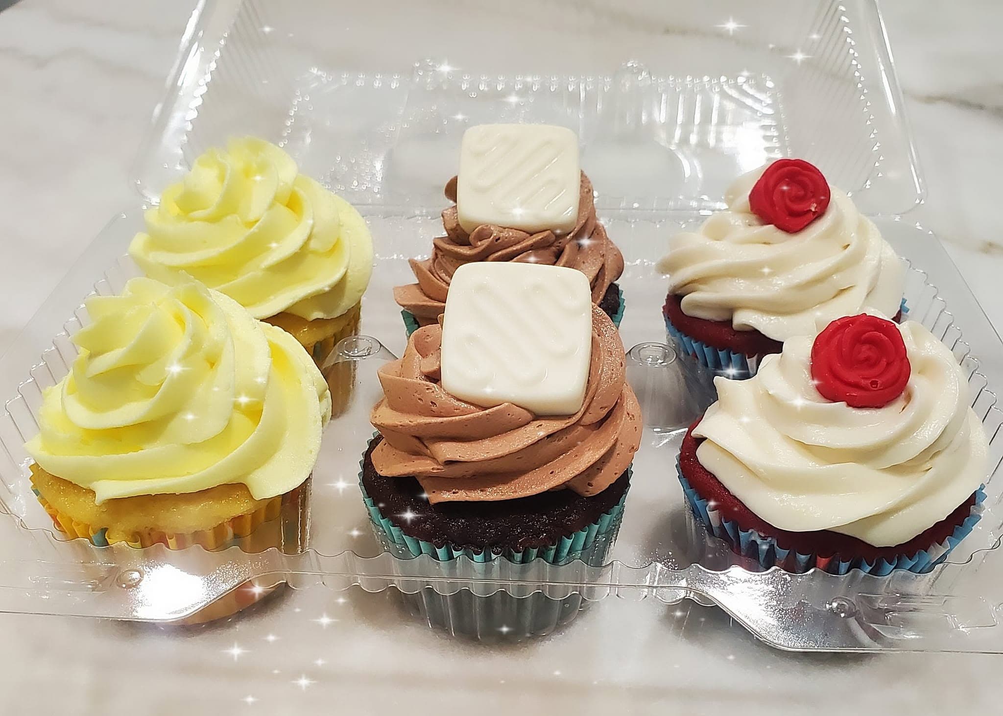 Six assorted cupcakes in a clear container with yellow, chocolate, and white frosting swirls and decorative toppings.