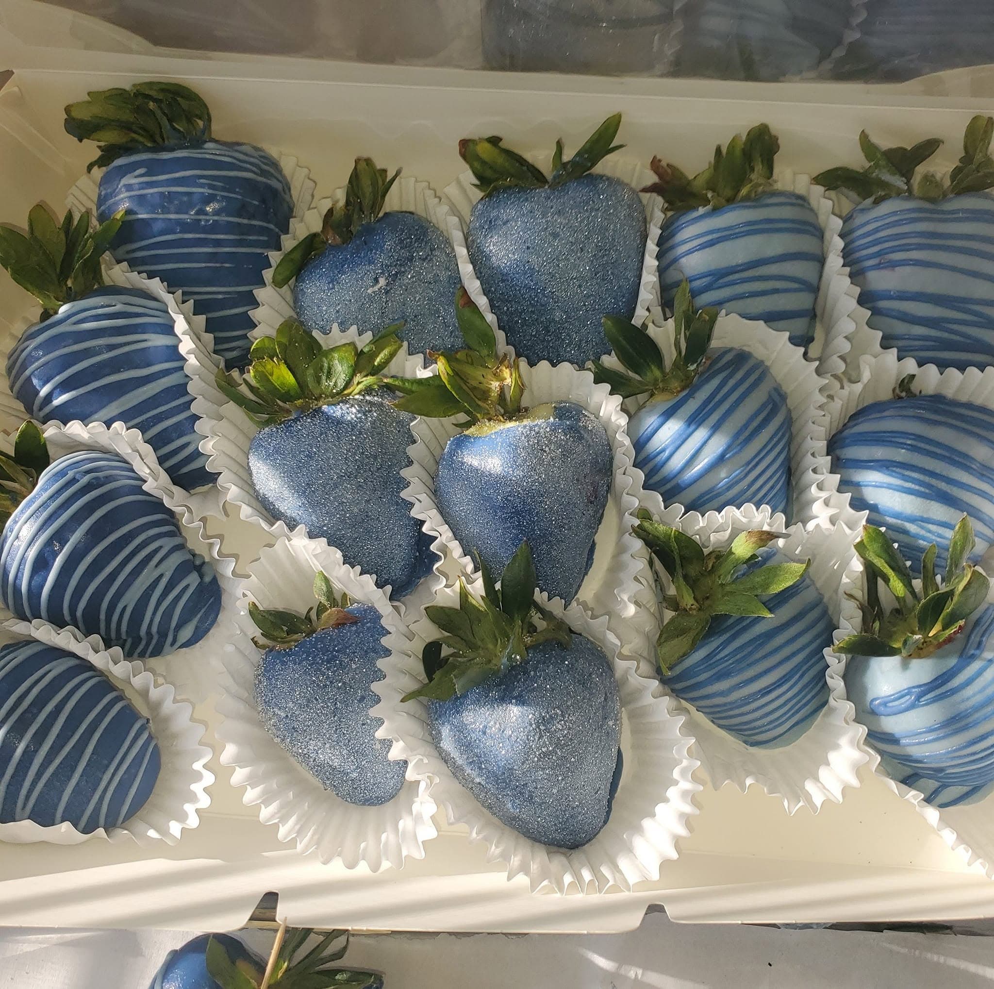 Blue chocolate-covered strawberries with glittery finishes and light blue stripes in white paper liners.