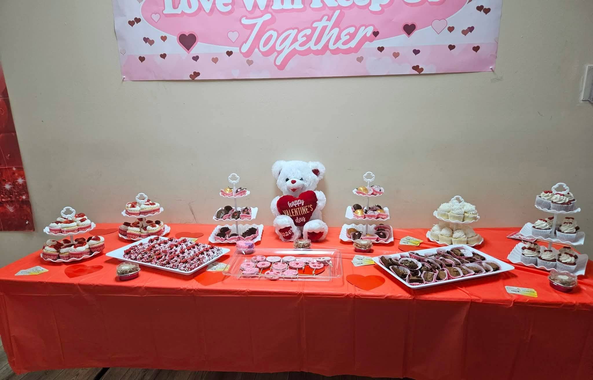 Valentine's Day dessert table with cupcakes, cookies, and a white teddy bear on red tablecloth.
