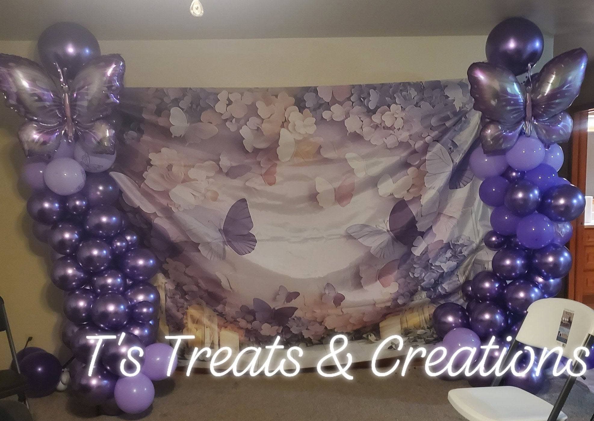 Purple butterfly-themed party backdrop featuring balloon columns and a floral butterfly print fabric background.