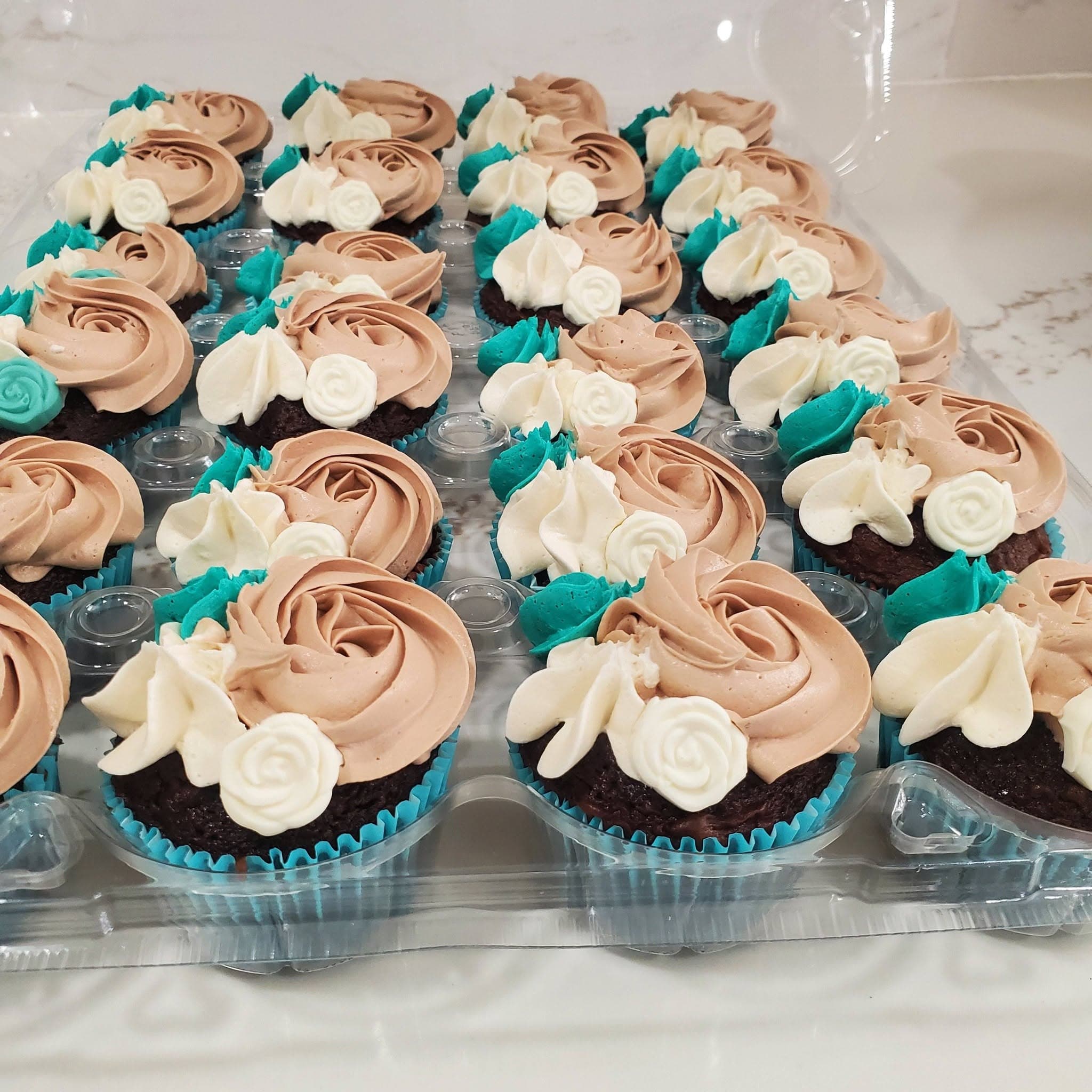 Chocolate cupcakes with mocha, white, and teal floral frosting swirls in a clear container.