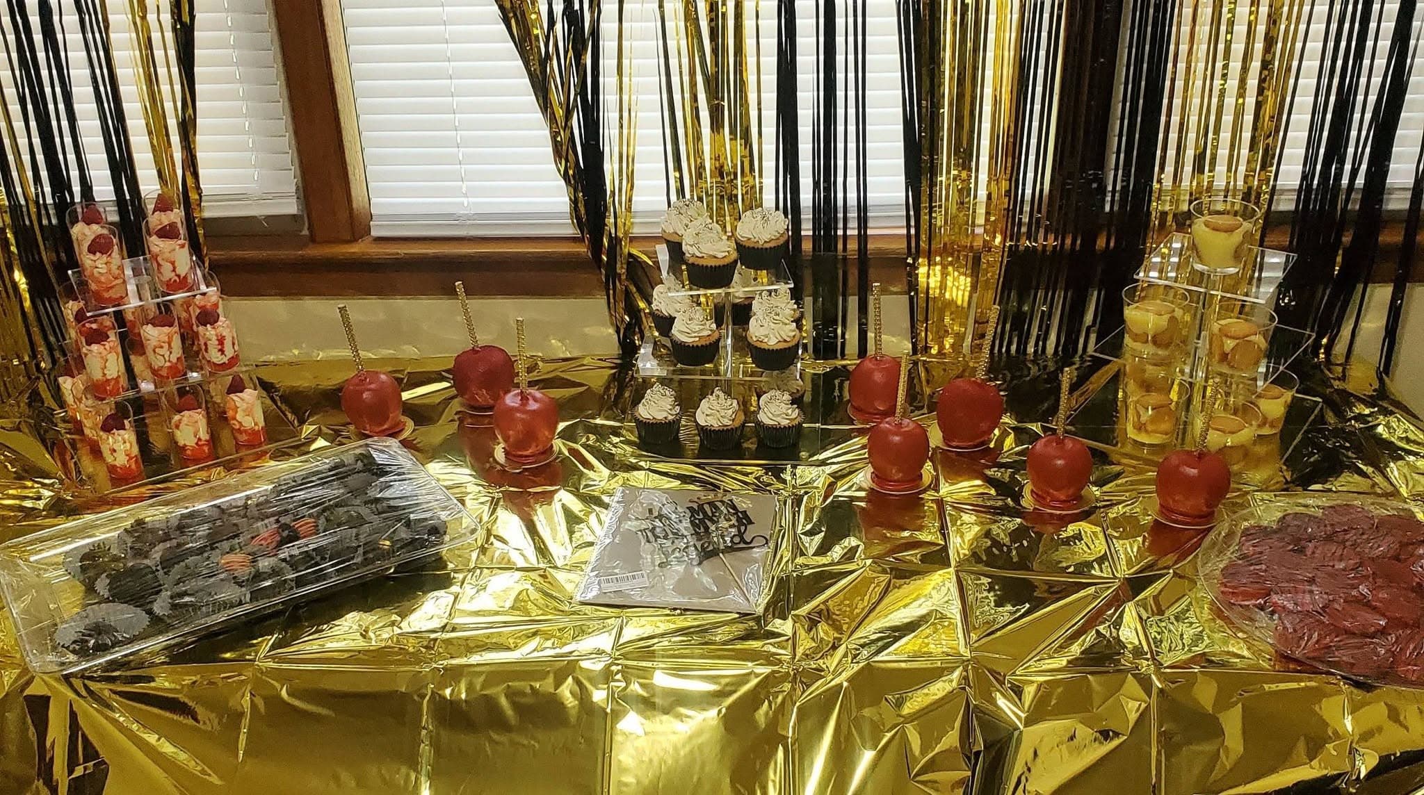 Gold foil table featuring red candy apples, cupcakes, and parfaits against black and gold tinsel.