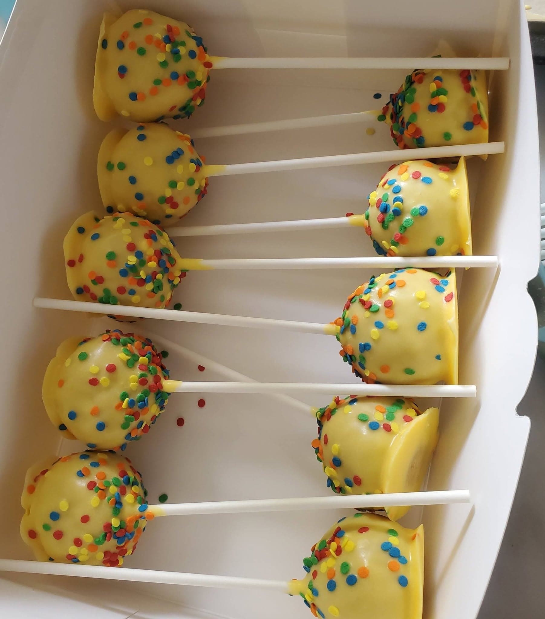 Yellow cake pops with colorful confetti sprinkles on white sticks arranged in a white box.
