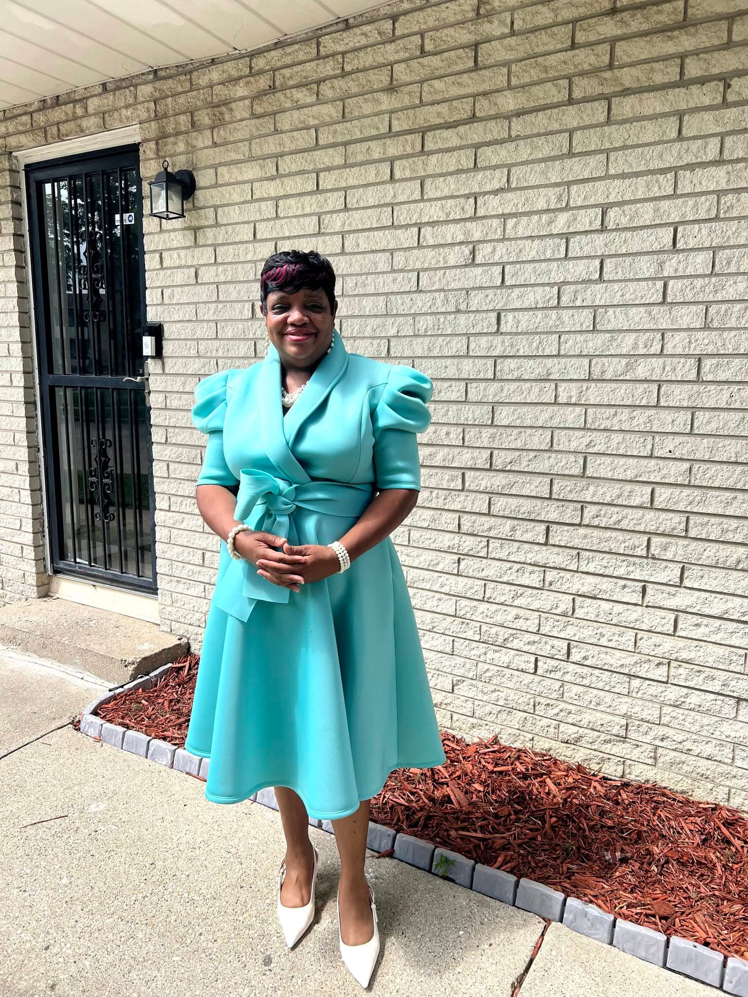 Smiling woman in a turquoise puff-sleeve dress and white heels stands before a brick wall.