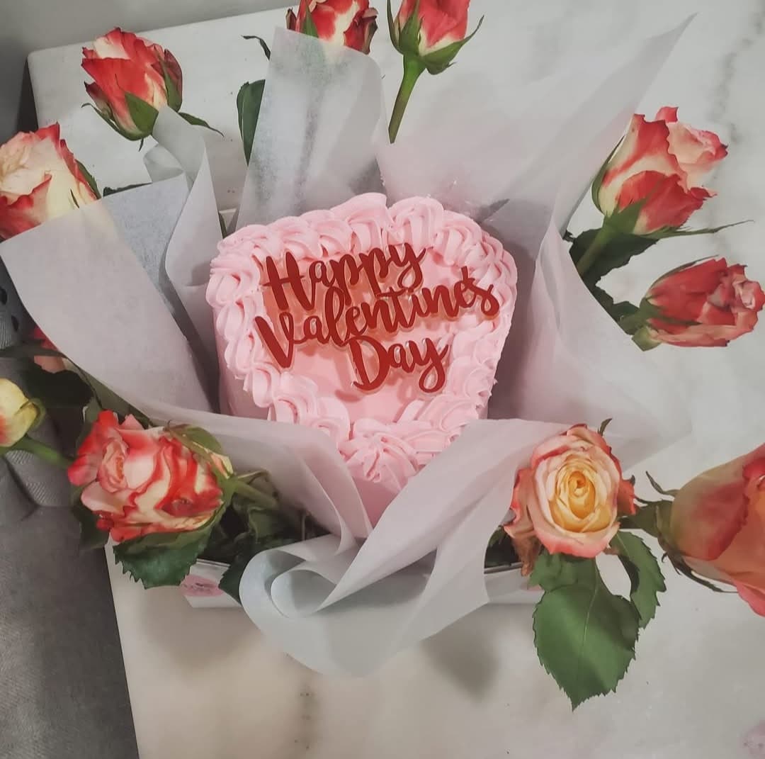 Pink heart cake with "Happy Valentine's Day" topper surrounded by red roses and white tissue.