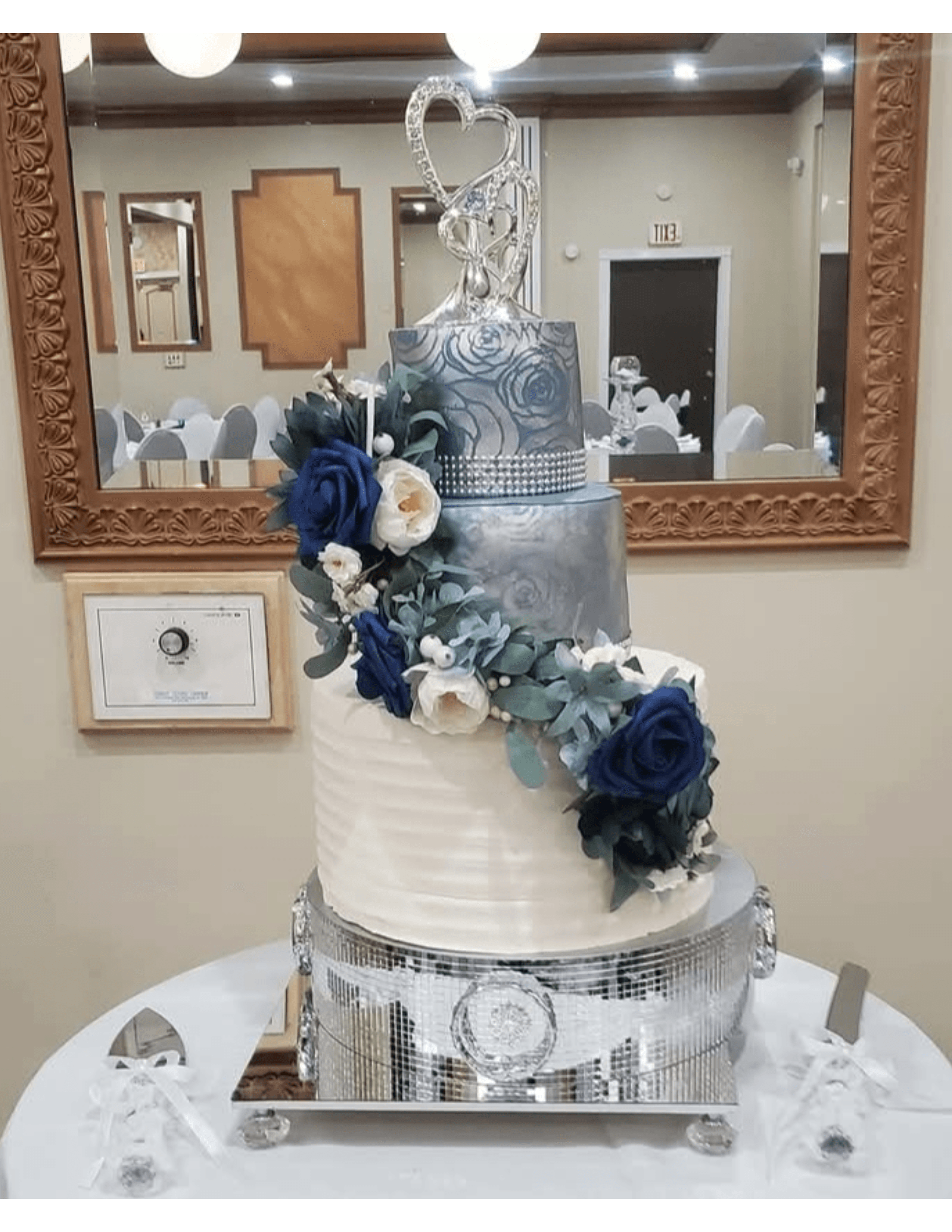 Three-tier silver and white wedding cake with blue floral cascade on a mirrored stand.