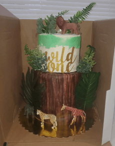 Two-tier jungle cake with tree trunk base, gold 'Wild One' script, and animal figurines.